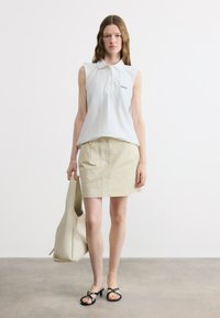 Γυναίκα που φοράει λευκό αμάνικο πόλο μπλουζάκι και μπεζ φούστα, κρατώντας μπεζ τσάντα ώμου και φοράει μαύρα πέδιλα με τακούνι ανοιχτά στα δάχτυλα, στέκεται σε γκρι πάτωμα.