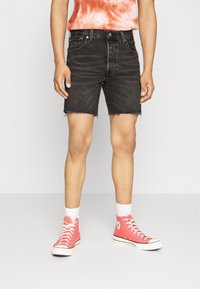 Pantalones cortos de mezclilla negra con dobladillo deshilachado, combinados con una camiseta de teñido anudado en naranja y blanco y zapatillas altas rosas. Calcetines blancos hasta el tobillo visibles.