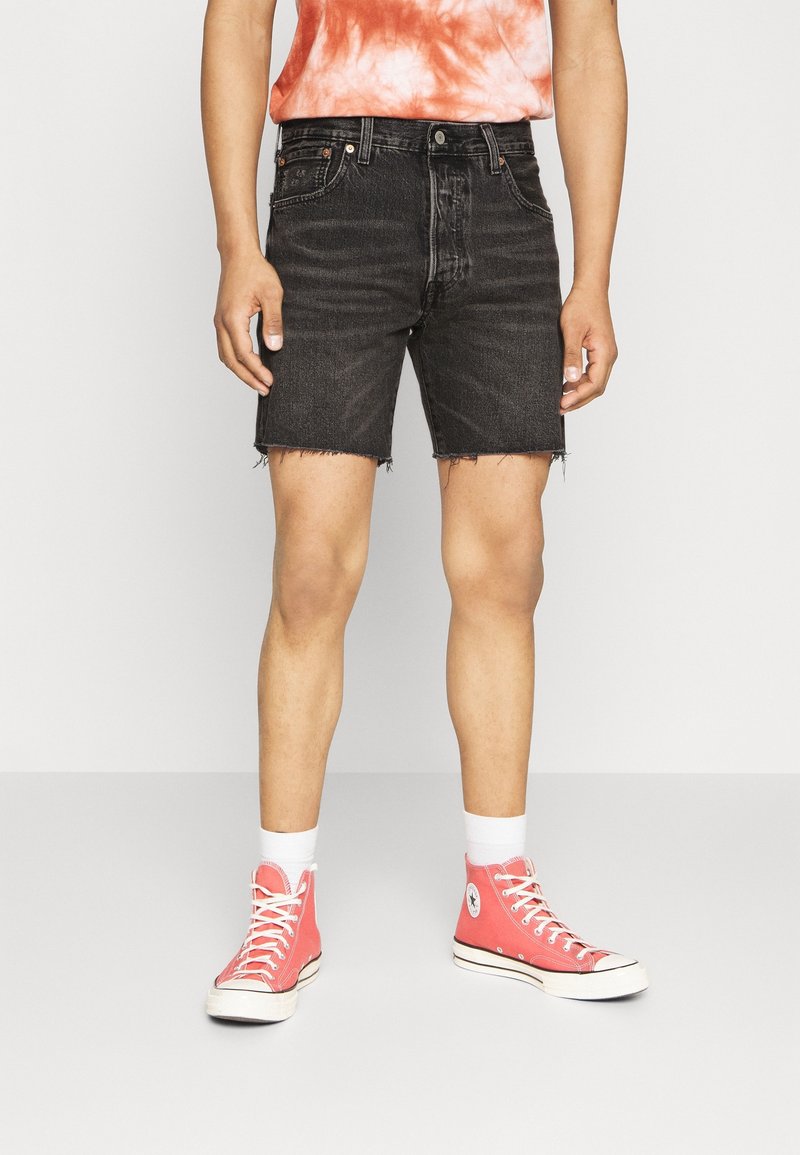 Pantalones cortos de mezclilla negra con dobladillo deshilachado, combinados con una camiseta de teñido anudado en naranja y blanco y zapatillas altas rosas. Calcetines blancos hasta el tobillo visibles.
