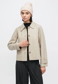 Beige ulljakke med krage, knapper foran og vide ermer. Kombinert med en svart topp og grå bukser.