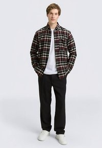 Plaid flannel shirt in burgundy and white, with a button-up design and chest pocket, worn over a white T-shirt and black pants.