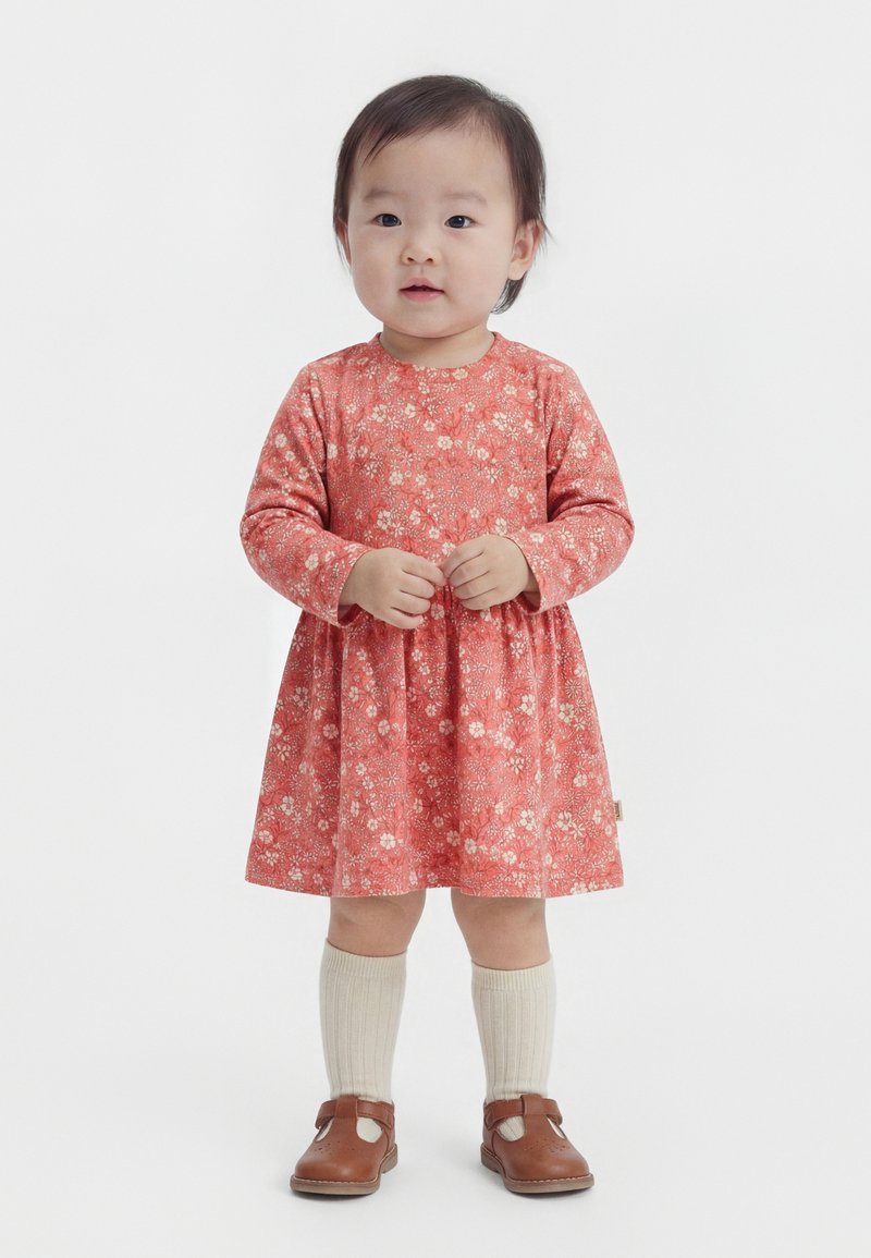 Copil mic în picioare, purtând o rochie roz cu flori și mânecă lungă, șosete crem până la genunchi și pantofi maro cu baretă, pe un fundal simplu.