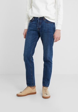 Person wearing blue straight-leg jeans, beige lace-up sneakers, and a white long-sleeve shirt standing against a plain background.