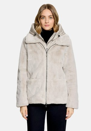 Femme portant une veste en fausse fourrure beige clair avec des poches avant et une capuche sur un haut noir, debout devant un fond clair uni.