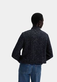 Giacca in tweed blu navy con particelle metalliche, design cropped, tessuto testurizzato e collo alto. Abbinata a jeans azzurri.