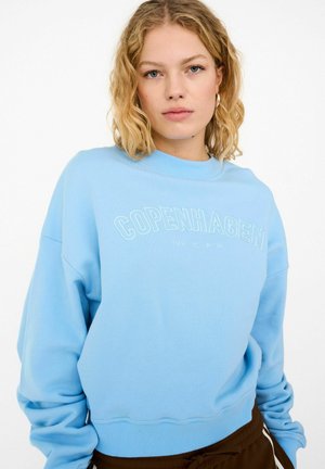 Blonde woman wearing light blue sweatshirt with "COPENHAGEN" text, standing against a plain white background.