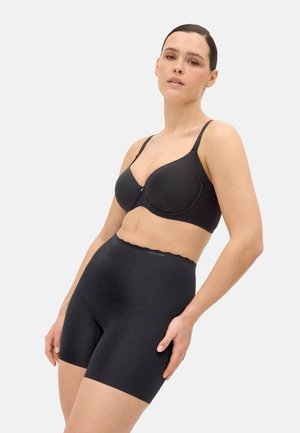 Mujer con shorts moldeadores negros sin costuras de cintura alta y sujetador negro con aro, posando contra un fondo claro liso.