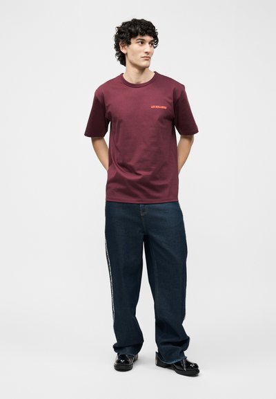 Νεαρός με σγουρά μαλλιά που φοράει μπορντό μπλουζάκι, σκούρα τζιν παντελόνια με φαρδιά μπατζάκια και πλαϊνές ρίγες και μαύρα παπούτσια, όρθιος με τα χέρια πίσω από την πλάτη.