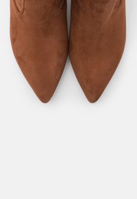 Bottes en daim marron à bout pointu avec une texture lisse, présentant des découpes décoratives sur les côtés et un design élégant.