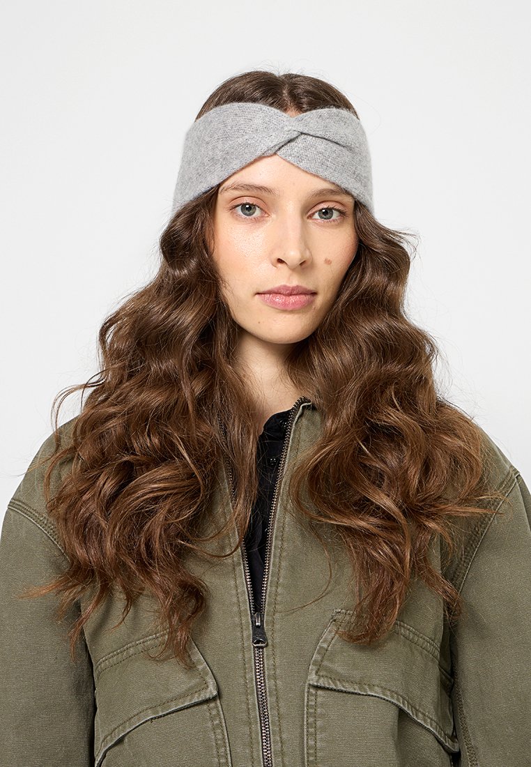 Grau gestricktes Wollhaarband mit gedrehtem Design, getragen von einer Person mit langen, gewellten Haaren. Der Hintergrund ist weiß, was das Accessoire hervorhebt.