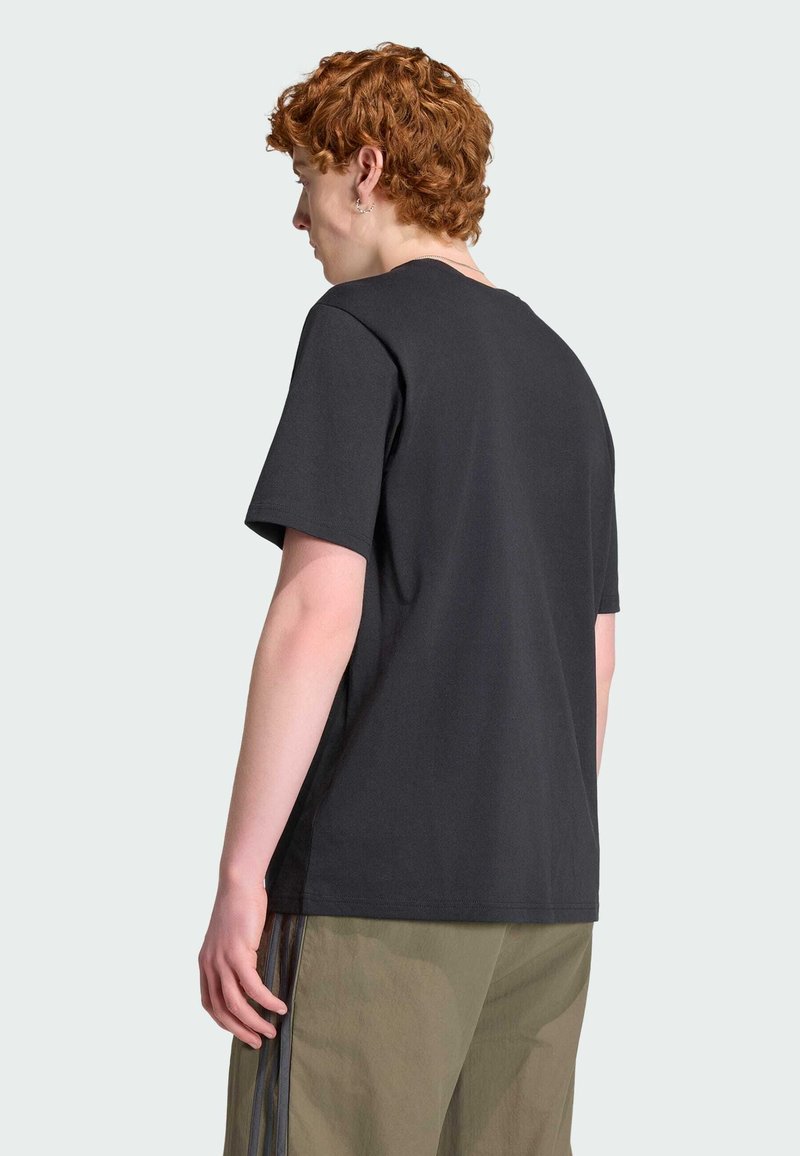 Jeune personne aux cheveux roux bouclés portant un t-shirt noir uni à manches courtes et un pantalon vert olive, vue de dos sur un fond clair.
