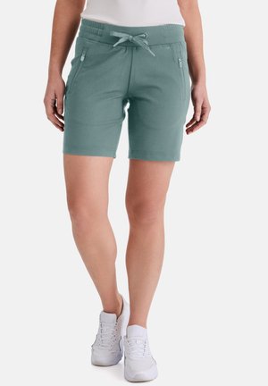 Person trägt salbeigrüne Shorts bis zur Mitte des Oberschenkels mit Kordelzugbund und Reißverschlusstaschen, kombiniert mit weißen Sportschuhen.
