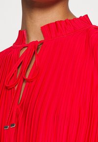 Rød plisseret bluse med en flæsekant, der har en nøglehulåbning og snoretræk med metalliske spidser. Glat stofstruktur.
