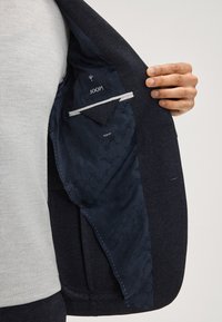 Marineblazer mit gemustertem dunkelblauem Futter. Verfügt über ein "JOOP!"-Label und eine Tasche, zeigt einen strukturierten Stoff und ein schlankes Design.