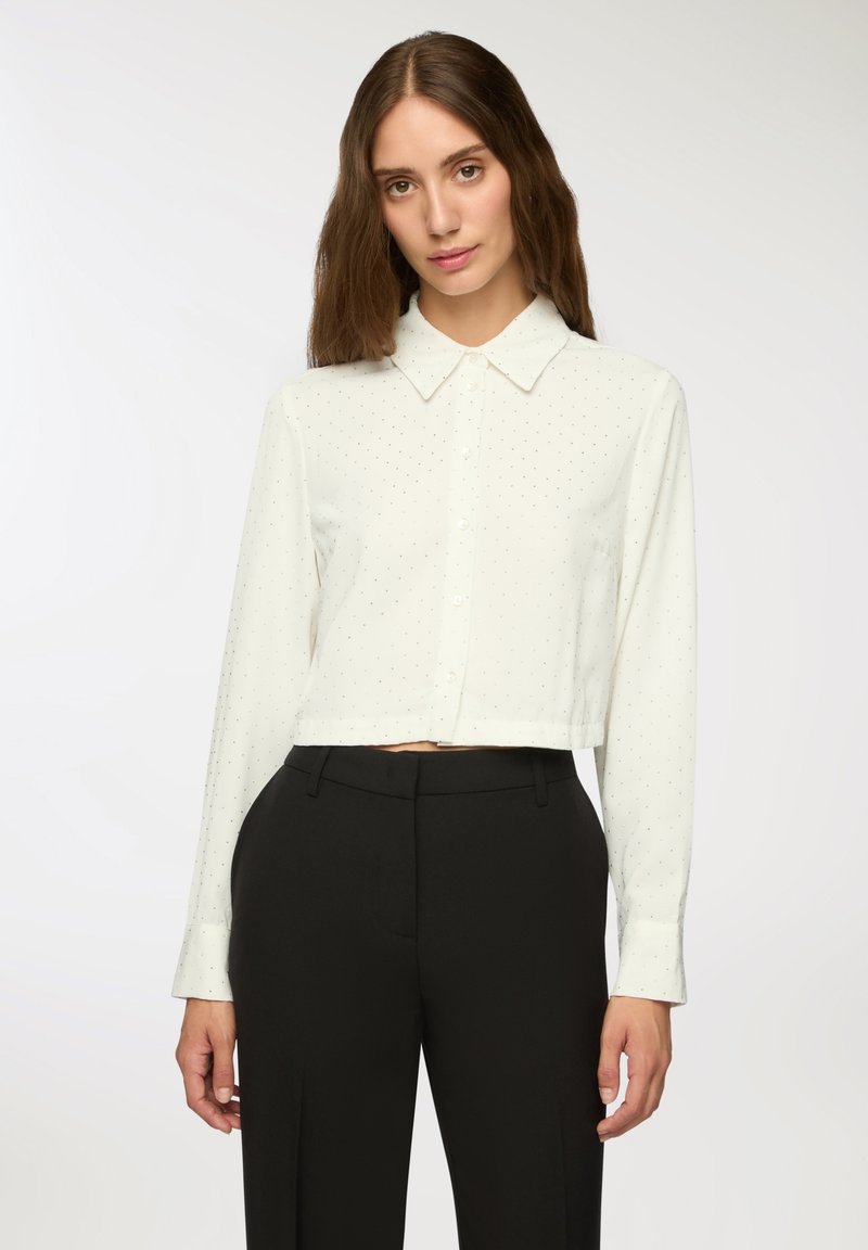 Camicia bianca a maniche lunghe con colletto button-down, caratterizzata da un sottile motivo a pois. Maniche lunghe con polsini bottonati. Abbinata a pantaloni neri.