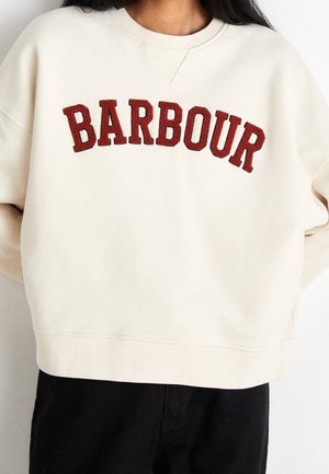 Bluza w kolorze kremowym z dużym czerwonym napisem "BARBOUR" na piersi, noszona przez osobę z długimi ciemnymi włosami.