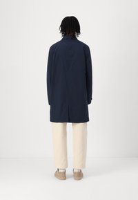 Persoon met donker gevlochten haar, gekleed in een marineblauwe lange jas, roomkleurige broek, witte sokken en beige schoenen, die voor een witte muur staat.