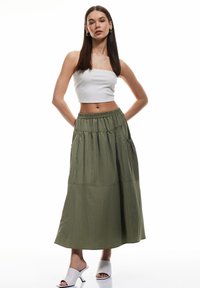 Maxi gonna verde oliva con vita elastica e tasche laterali, abbinata a un top corto bianco aderente e sandali con tacco grigio chiaro.