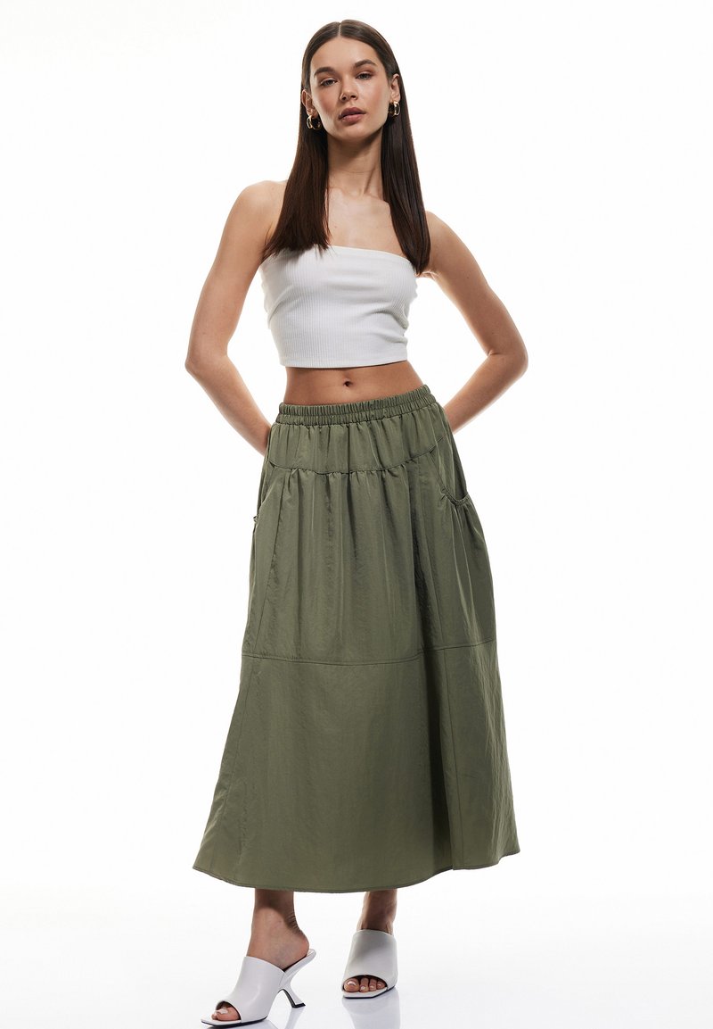 Maxi gonna verde oliva con vita elastica e tasche laterali, abbinata a un top corto bianco aderente e sandali con tacco grigio chiaro.