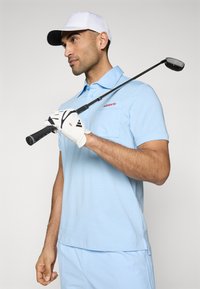 Light blue polo shirt with a pocket and red logo, worn with a white cap. Holding a golf club with a white glove featuring black accents.