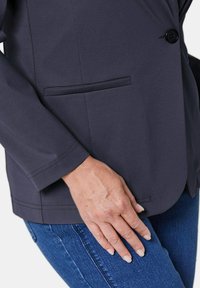 Donkerblauwe blazer van gladde stof, met een enkele knoop, één zak aan de linkerkant en gedetailleerde stiksels aan de randen.