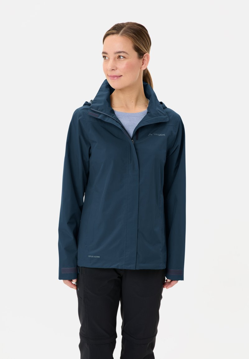 Veste marine imperméable avec capuche et fermeture éclair, présentant une texture lisse et un design sportif, associée à un pantalon noir.