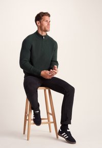 Knitted green sweater with high collar and zip, worn with black jeans and black sneakers, seated on a wooden stool against a light background.