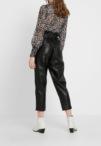 Svart läderbyxor med hög midja och avslappnad passform, i kombination med en genomskinlig mönstrad blus i svart och vitt samt vita ankelskór.