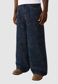 Ciemnoniebieskie, szerokie dżinsy z wzorem ciągłej pomarańczowej linii. Tkanina ma teksturowany wygląd, uzupełniony widocznym przeszyciem.