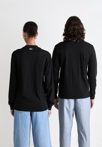 Zwei Personen stehen mit dem Rücken zueinander, tragen schwarze Langarmshirts mit grünen Krokodillogos auf dem oberen Rücken und hellfarbene Hosen.
