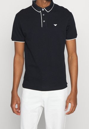 Mand iført en sort kortærmet poloshirt med hvide kanter og logo, kombineret med hvide bukser, stående mod en ensfarvet baggrund.