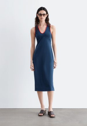 Femme portant une robe midi bleu marine sans manches avec un liseré rouge, des sandales marron à talons ouverts, des lunettes de soleil et des bracelets argentés, debout sur un sol gris.