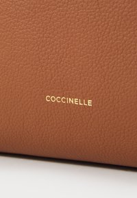 Braun genarbtes Leder mit eingeprägtem "COCCINELLE" in Gold. Nahaufnahme-Details betonen die Struktur und Farbe des Materials.