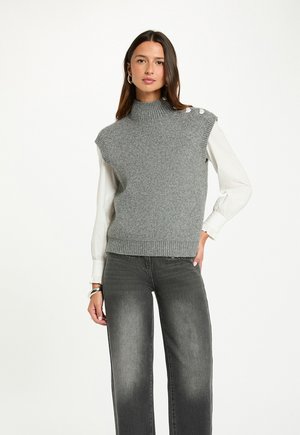 Gilet a costine grigio con alto collo e dettagli con bottoni sulle spalle, indossato sopra una camicia bianca a maniche lunghe. Abbinato a jeans grigi a gamba larga.