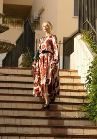 Maxiklänning i röd och krämfärgad med blommönster, långa ärmar, insnörd midja, parat med svarta sandaletter, nedstiga stentrappor.