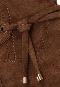 Primer plano de una bota de ante marrón que presenta un cordón anudado con puntas metálicas, costuras visibles y un detalle de ojal redondeado.