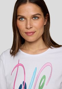 White shirt featuring multicolored rhinestone lettering in pink, blue, green, and dark blue. Classic crew neckline and short sleeves.