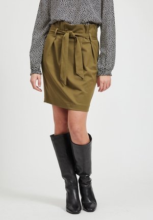 Vrouw draagt een olijfgroene rok met hoge taille met strik aan de voorkant, een blouse met lange mouwen met patroon en zwarte leren kniehoge laarzen.