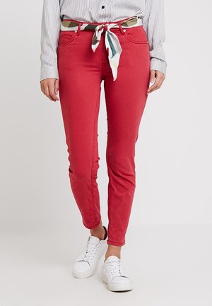 Frau trägt rote Slim-Fit-Hose mit mehrfarbigem, gebundenem Schal als Gürtel, weiße Turnschuhe und ein gestreiftes, weißes Hemd zum Knöpfen.