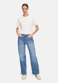 Hvid bomulds t-shirt med grøn tekst "oh la la" parret med lyseblå vide jeans og beige ankelstøvler. Modellen står med hænderne i lommerne.