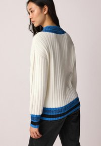 Weißer gerippter Pullover mit blauem Strickkragen und kontrastierenden blauen und schwarzen Streifen am Saum und an den Bündchen. Lockere Passform, lange Ärmel.