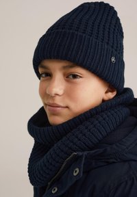 Temno modra pletena kapa z rebrastim vzorcem, v kompletu z ujemajočim se rebrastim šalom. Mehak material in prilegan oblikovanje za toploto.