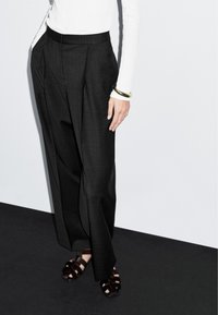 Zwarte geplooide broek met een gestructureerde afwerking, gecombineerd met donkere open teensandalen. Het model draagt een witte top met lange mouwen en een gouden armband.