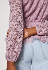 Lavendelfärgad spetsblus med strukturerat blommönster, elastiska puffärmar och en scalloped fåll, kombinerad med ljusblå höga jeans.