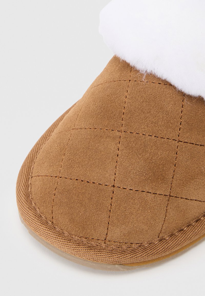 Braune Wildlederhausschuhe mit einem gesteppten Muster, einer weichen weißen Innenausstattung und einer beige Gummisohle, versehen mit genähten Details an den Rändern.