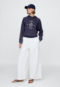 Tengerészkék kapucnis pulóver logóval, fehér, bőszárú nadrág és fekete papucs. Pamut anyag, laza fazon, látható varrások és gombos részletek.