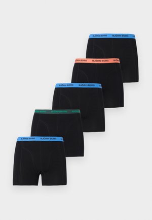 Νέος άνδρας ξαπλωμένος στο πλάι πάνω σε χαλί, φοράει μαύρα μποξεράκια Björn Borg, κοιτάζει σκεπτικός προς τα δεξιά.