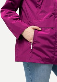 Fuchsia jacka med en glänsande, slät textur; har en ficka med dragkedja och ett elastiskt midjeband; kombinerad med ljusblå jeans.