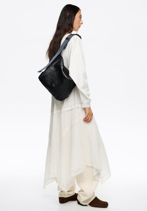 Czarna skórzana torba w stylu slouchy z niebieskim regulowanym paskiem, z delikatnym logo i gładką teksturą, noszona na białej, warstwowej sukience.