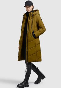 Abrigo largo acolchado en verde oliva con capucha, cierre de cremallera y cordones. Acentos en negro y bolsillos con textura suave. Lleva puesto sobre pantalones negros y botas.